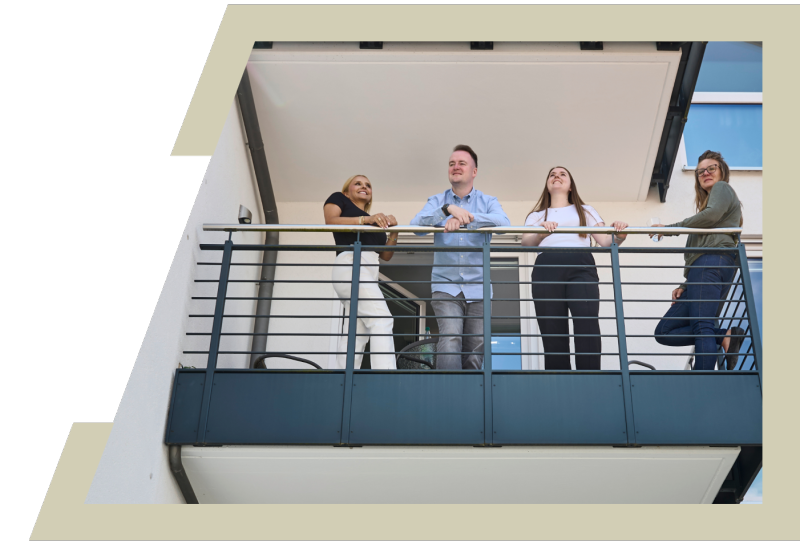Foto: Das Bild zeigt vier Personen, die gemeinsam auf einem modernen Balkon eines Gebäudes stehen. Sie lehnen sich locker an das Balkongeländer und schauen nach vorne. Die Gruppe besteht aus zwei Frauen links und rechts außen, einem Mann und einer weiteren Frau in der Mitte. Der Hintergrund zeigt die Fassade des Hauses mit Fenstern und heller Wandfarbe. Das Foto vermittelt eine offene, freundliche und entspannte Stimmung. - 